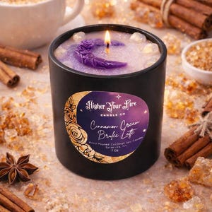 Op de afbeelding: Een zwarte kaars met een brandende lont, met een paarse maan en bloemenontwerp. Het etiket luidt "Higher Your Fire Candle Co" en "Cinnamon Cream Brulee Latte". Kaneelstokjes en een kopje koffie staan op de achtergrond.