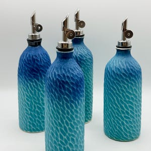 May include: Four cylindrical glass bottles with silver-colored metal pour spouts. Each bottle features a textured, hammered surface with a blue ombre gradient, transitioning from a deep navy blue at the top to a bright turquoise at the base.