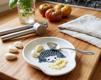 Puede incluir: Un plato de ajo de cerámica blanca con un diseño de erizo azul, una cuchara de acero inoxidable y un prensador de ajos se encuentran sobre una tabla de cortar de madera. También hay dientes de ajo frescos y ajo picado. Tomates y hierbas en el fondo.