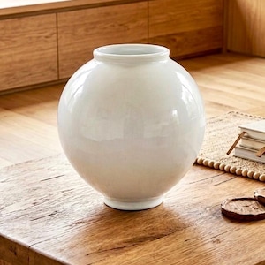 May include: A white, spherical ceramic vase with a wide base and a rounded top. The vase sits on a wooden table with visible wood grain. The background features wooden cabinetry and flooring, creating a neutral, minimalist aesthetic.