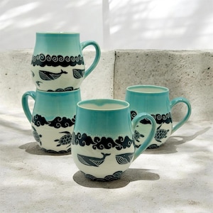 May include: Four ceramic mugs with a teal glaze and white base. Each mug features a black wave design with whales, turtles, and leaf illustrations. The mugs have a curved handle and a smooth, glossy finish.