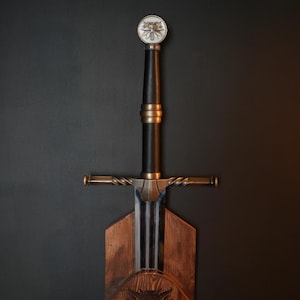 May include: A longsword with a black grip, a metallic crossguard with a twisted design, and a circular pommel featuring a wolf head emblem. The sword is mounted on a wooden plaque with a dark finish, displaying a carved wolf head symbol.