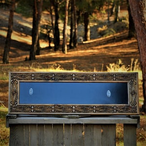 Könnte beinhalten: Eine rechteckige Holzvitrine mit dunkelblauem Innenraum und aufwendigen Schnitzereien. Der Rahmen ist warmbraun und die Vitrine hat zwei silberfarbene Metallverschlüsse. Die Vitrine steht auf einer Holzoberfläche mit verschwommenem Waldboden.