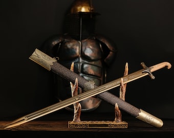 May include: An antique sword with a gold blade and a wooden handle rests in a decorative stand. The scabbard is dark brown with gold accents. A suit of armor and helmet are in the background.