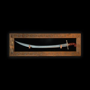 Puede incluir: Una espada plateada con mango rojo y dorado se exhibe en un marco de madera con tallas intrincadas. La espada tiene caligraf&iacute;a &aacute;rabe en la hoja.
