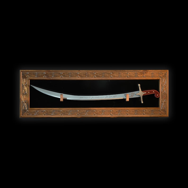 Puede incluir: Una espada plateada con mango rojo y dorado se exhibe en un marco de madera con tallas intrincadas. La espada tiene caligraf&iacute;a &aacute;rabe en la hoja.