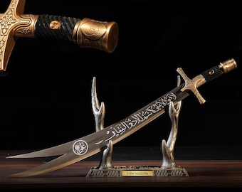 May include: A decorative sword with a curved blade, displayed on a stand. The sword features a gold-colored hilt and crossguard, with intricate detailing. The handle is wrapped in black material. The blade has engraved text and a circular emblem.