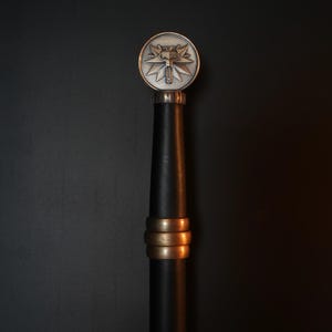 May include: A sword hilt featuring a circular silver-toned pommel with a raised wolf head emblem. The grip is wrapped in black material and accented with three horizontal bronze-toned rings positioned below the pommel and above the handle base.