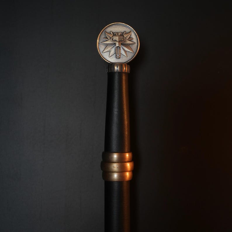 May include: A sword hilt featuring a circular silver-toned pommel with a raised wolf head emblem. The grip is wrapped in black material and accented with three horizontal bronze-toned rings positioned below the pommel and above the handle base.
