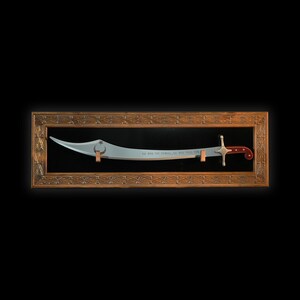 May include: A silver-colored scimitar sword with a red and silver handle is displayed in a wooden frame with intricate carvings. The sword has a crescent moon and star design on the blade and the inscription "As God is the witness, so shall justice be done".
