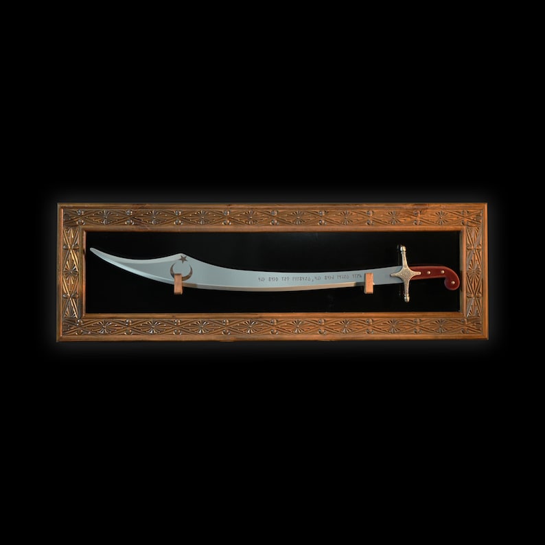 May include: A silver-colored scimitar sword with a red and silver handle is displayed in a wooden frame with intricate carvings. The sword has a crescent moon and star design on the blade and the inscription "As God is the witness, so shall justice be done".