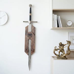 May include: A silver-colored metal sword with a black grip and a circular pommel, mounted vertically on a dark brown wooden wall plaque. The plaque features a black wolf head emblem engraved in the center, holding the sword in place.