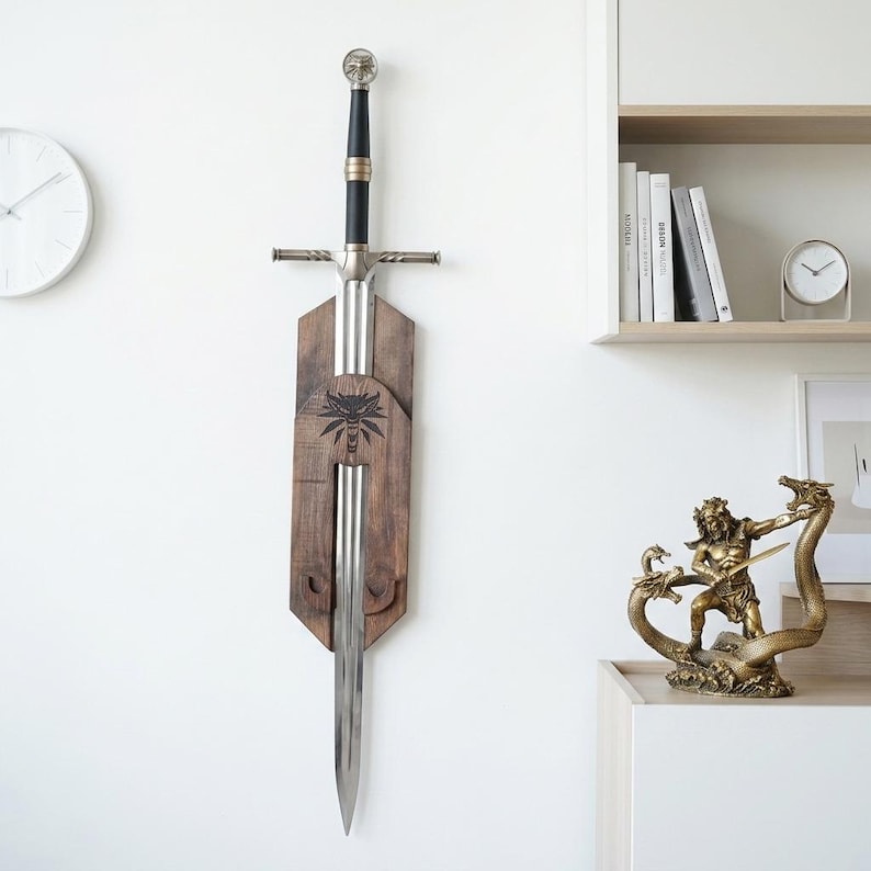 May include: A silver-colored metal sword with a black grip and a circular pommel, mounted vertically on a dark brown wooden wall plaque. The plaque features a black wolf head emblem engraved in the center, holding the sword in place.