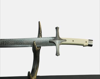 Pode incluir: Uma espada prateada com uma pega branca e uma guarda em forma de cruz. A espada é exibida num suporte de madeira.