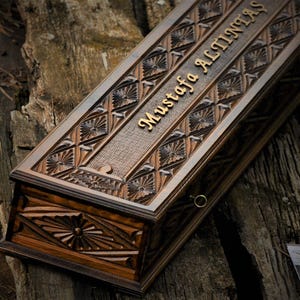 Könnte beinhalten: Eine rechteckige Holzkiste mit aufwendig geschnitzten floralen und geometrischen Mustern. Die Kiste ist dunkelbraun mit goldfarbenem Schriftzug "Mustafa Altintas". An der Seite ist ein kleiner Metallring zu sehen.