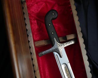 Puede incluir: Una espada con mango de madera oscura con tres remaches metálicos, una guarda de plata con grabados florales intrincados y una hoja de acero con inscripciones doradas, dentro de un estuche de madera forrado con tela de terciopelo rojo.