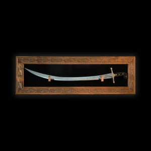Puede incluir: Una espada plateada con mango negro se muestra en un marco de madera con un dise&ntilde;o ornamentado. La espada tiene escritura &aacute;rabe en la hoja.