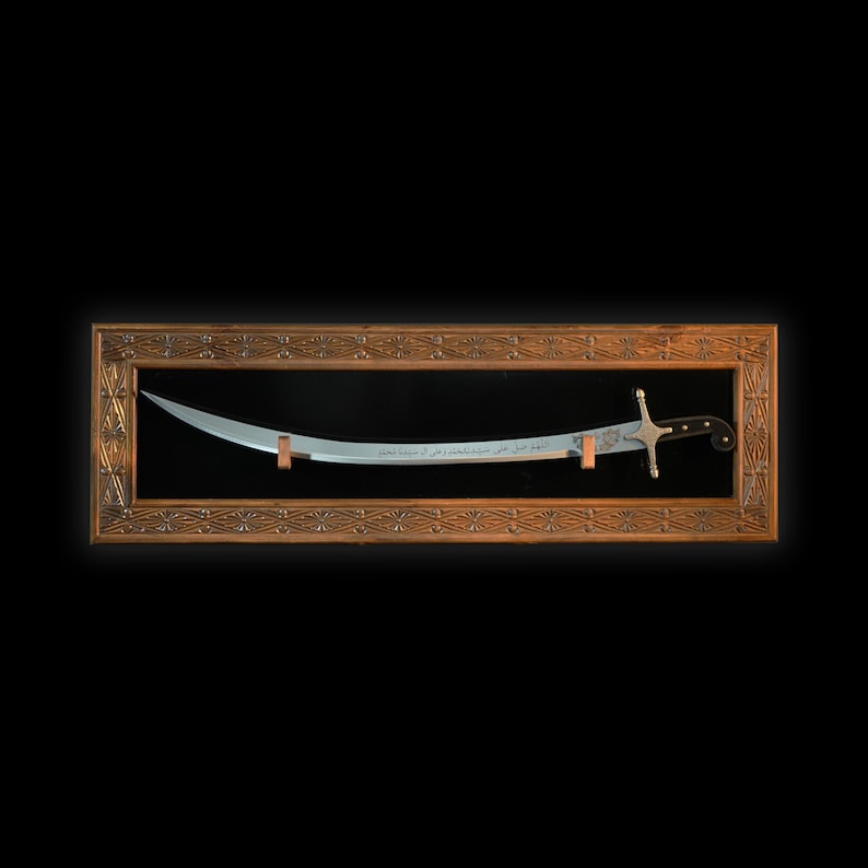 Puede incluir: Una espada plateada con mango negro se muestra en un marco de madera con un dise&ntilde;o ornamentado. La espada tiene escritura &aacute;rabe en la hoja.