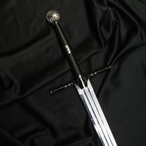 May include: A silver-toned metal sword featuring a long, double-edged blade and a black cylindrical handle. The crossguard has a twisted metal design, and the pommel is topped with a circular medallion displaying a detailed wolf head emblem.