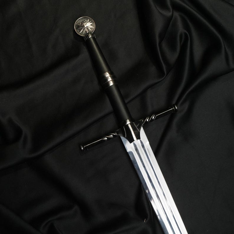 May include: A silver-toned metal sword featuring a long, double-edged blade and a black cylindrical handle. The crossguard has a twisted metal design, and the pommel is topped with a circular medallion displaying a detailed wolf head emblem.
