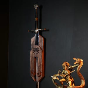 May include: A long steel sword with a black grip and a circular pommel, mounted vertically on a dark brown wooden wall plaque. The plaque features a carved wolf emblem in the center. A gold-colored statue of a figure fighting a multi-headed serpent.