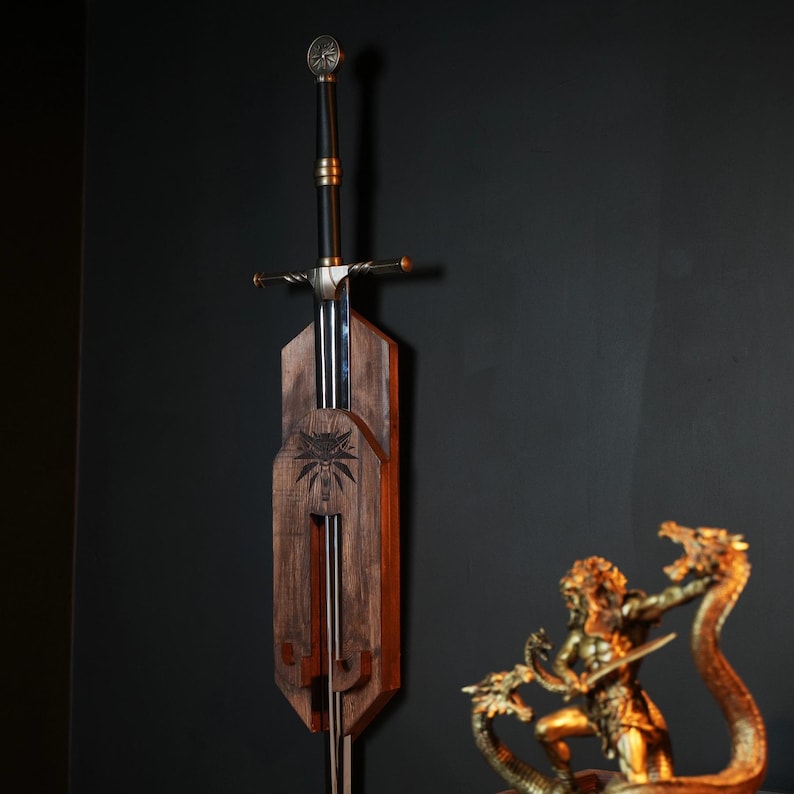 May include: A long steel sword with a black grip and a circular pommel, mounted vertically on a dark brown wooden wall plaque. The plaque features a carved wolf emblem in the center. A gold-colored statue of a figure fighting a multi-headed serpent.