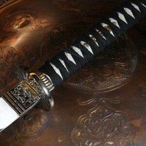 Peut inclure: Gros plan sur un katana. La poign&eacute;e est envelopp&eacute;e de tissu noir avec des accents blancs en forme de losange. La garde est argent&eacute;e avec des motifs d&eacute;coup&eacute;s complexes. La lame est blanche et l'&eacute;p&eacute;e repose sur une surface marron &agrave; motifs.