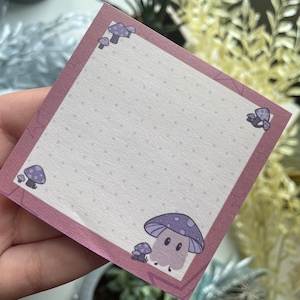 May include: A pink notepad with a white background and a purple mushroom design. The notepad has a dotted pattern and a border of purple mushrooms.