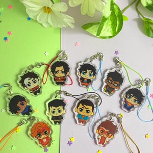 May include: A selection of cartoon character acrylic charms featuring superhero designs. Each clear charm has a colourful character illustration, attached to a silver keychain and a coloured lanyard. The background is a mix of green and white with star-shaped confetti.