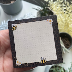 以下が含まれることがあります： ハニカムの縁取りとドット柄の黒と白の蜂をテーマにしたメモ帳。メモ帳には蜂が2匹と花がいくつか描かれています。