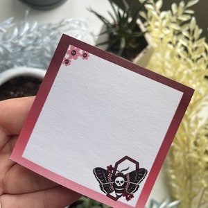 May include: A burgundy and white card with a black and white moth with a skull design. The moth is inside a hexagon with flowers. The card has a pink border with small pink flowers in the corner.