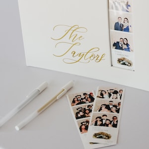 May include: A cream-coloured photo album with gold script text reading "The Taylors" and a window showing photo booth strips. Two pens and extra photo strips are placed beside the album. The album is open.