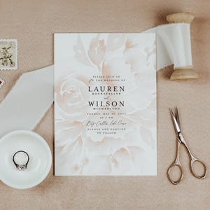 Op de afbeelding: Trouwkaart met bloemenontwerp, met de namen Lauren Rockefeller en Wilson Michaelson. Inclusief een ring in een wit schaaltje, vintage postzegels, wit lint, een schaar en een houten spoel, alles op een neutrale achtergrond.