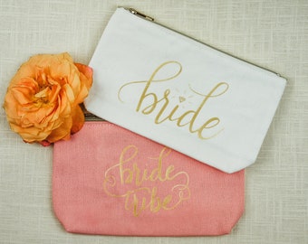 May include: Two white and pink canvas zippered pouches with gold lettering. The white pouch says "bride" with a diamond symbol. The pink pouch says "bride tribe".
