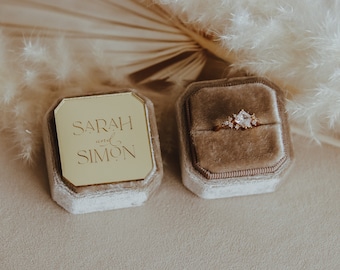 May include: A gold ring box with a velvet interior. The box is open and a ring with a large, clear stone and smaller stones is inside. The box lid has the inscription "SARAH and SIMON" in gold lettering.