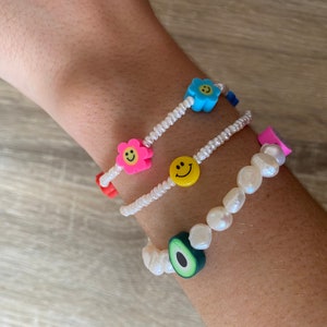 May include: Three beaded bracelets with colourful charms. The first bracelet has a blue flower charm, the second has a pink flower charm and a yellow smiley face charm, and the third has a green avocado charm.