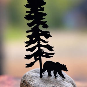 May include: A black metal silhouette of a tall pine tree and a walking bear mounted onto a smooth, light grey, oval-shaped river stone. The metal cutout features detailed branches on the tree and the distinct shape of a bear in profile.