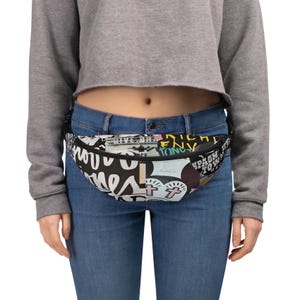 May include: A black fanny pack with a colorful, graphic print of various words and images. The fanny pack is worn around the waist of a person wearing a grey sweater and blue jeans.