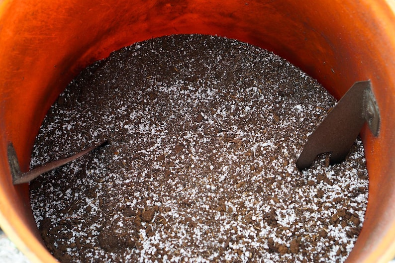 K&ouml;nnte beinhalten: Ein orangefarbener Beh&auml;lter gef&uuml;llt mit dunkelbrauner Erde und wei&szlig;em Perlit. Zwei braune Spaten sind teilweise sichtbar, was auf Garten- oder Pflanzaktivit&auml;ten hindeutet. Die Erde scheint bereit zum Pflanzen zu sein.