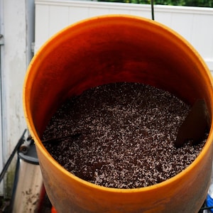 K&ouml;nnte beinhalten: Ein orangefarbener Kunststoffbeh&auml;lter gef&uuml;llt mit dunkelbrauner Erde und wei&szlig;en Granulaten. Eine braune Kelle ist teilweise im Inneren des Beh&auml;lters zu sehen. Der Beh&auml;lter befindet sich im Freien, mit einer wei&szlig;en T&uuml;r und einem Zaun im Hintergrund.