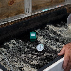 A worm bin that is being monitored for temperature and moisture. A hand is applying worm feed and grit to the worm bin showcasing the diet the worms receive.