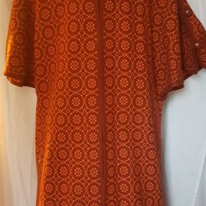 Op de afbeelding: Een lange, oranje en bruine gebreide cardigan met een ritssluiting. De cardigan heeft een geometrisch patroon en korte mouwen.