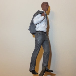 May include: A grey suit jacket is draped over the shoulder of a figurine wearing a white dress shirt and grey trousers. The figurine is standing with one leg slightly bent and the other leg straight.