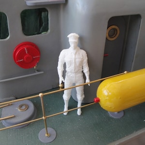 May include: A white plastic model of a sailor standing on a grey ship deck. The ship has a red fire hose reel and a yellow torpedo. The sailor is wearing a white uniform and a black cap.