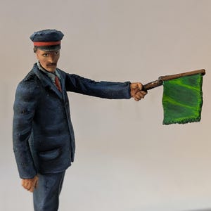 May include: A 3D printed figurine of a railway conductor wearing a blue uniform and a cap with a red band. The conductor is holding a green flag with a wooden stick.