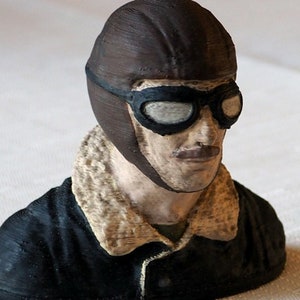 May include: A 3D printed bust of a pilot wearing a brown leather helmet with goggles and a black jacket with a brown fur collar.