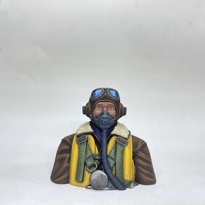 May include: A detailed bust of a World War II pilot. The figure wears a brown leather jacket with a yellow vest, a blue oxygen mask, and brown goggles. The pilot's helmet is brown with blue lenses.