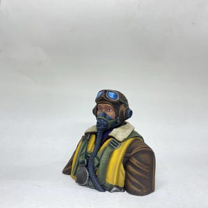 May include: A detailed bust of a World War II pilot. The figure wears a brown leather aviator helmet with blue-tinted goggles, a yellow jacket, and a dark blue oxygen mask. The bust is painted with realistic details.