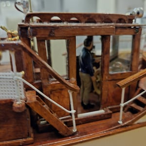 May include: A detailed wooden model of a boat with a white railing and a small figure of a person standing in the cabin. The cabin has a glass window and a wooden door.
