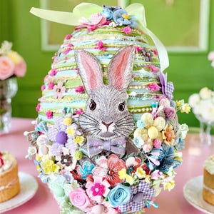 Può includere: Decorazione a forma di uovo di Pasqua con un coniglio grigio, orecchie rosa e un papillon viola. L'uovo è decorato con fiori e nastri colorati nei toni del rosa, blu e giallo, su sfondo verde.